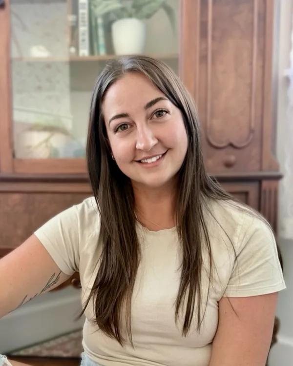 Ashley Tipple, owner, smiling, seated in front of a wooden china cabinet.
