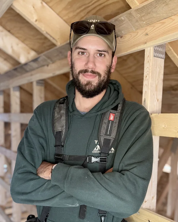 Bradley Tipple, owner and carpenter, arms crossed in a framed attic, sunglasses on his company cap and his tool belt on.