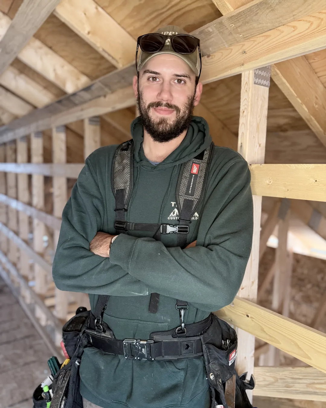Bradley Tipple, owner, arms crossed in the framing of an attic, in his green company hoodie and cap with his tool harness on.