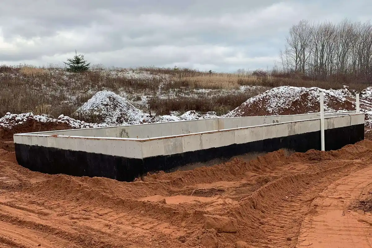 A poured and waterproofed foundation sitting in cut red earth, first snow across the spoil banks behind it.